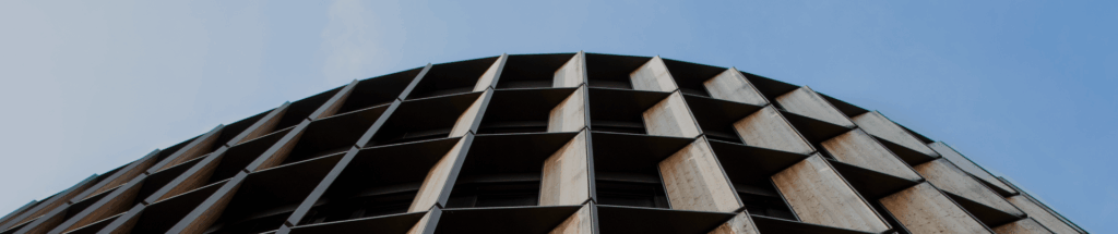 Upward view of curved brutalist building facade with distinctive diamond-shaped concrete panels creating a dramatic geometric pattern against a clear pale blue sky.